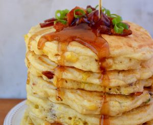 Tall stack of Sweet Corn Pancakes with Bacon topped with sliced scallions and drizzled with maple syrup